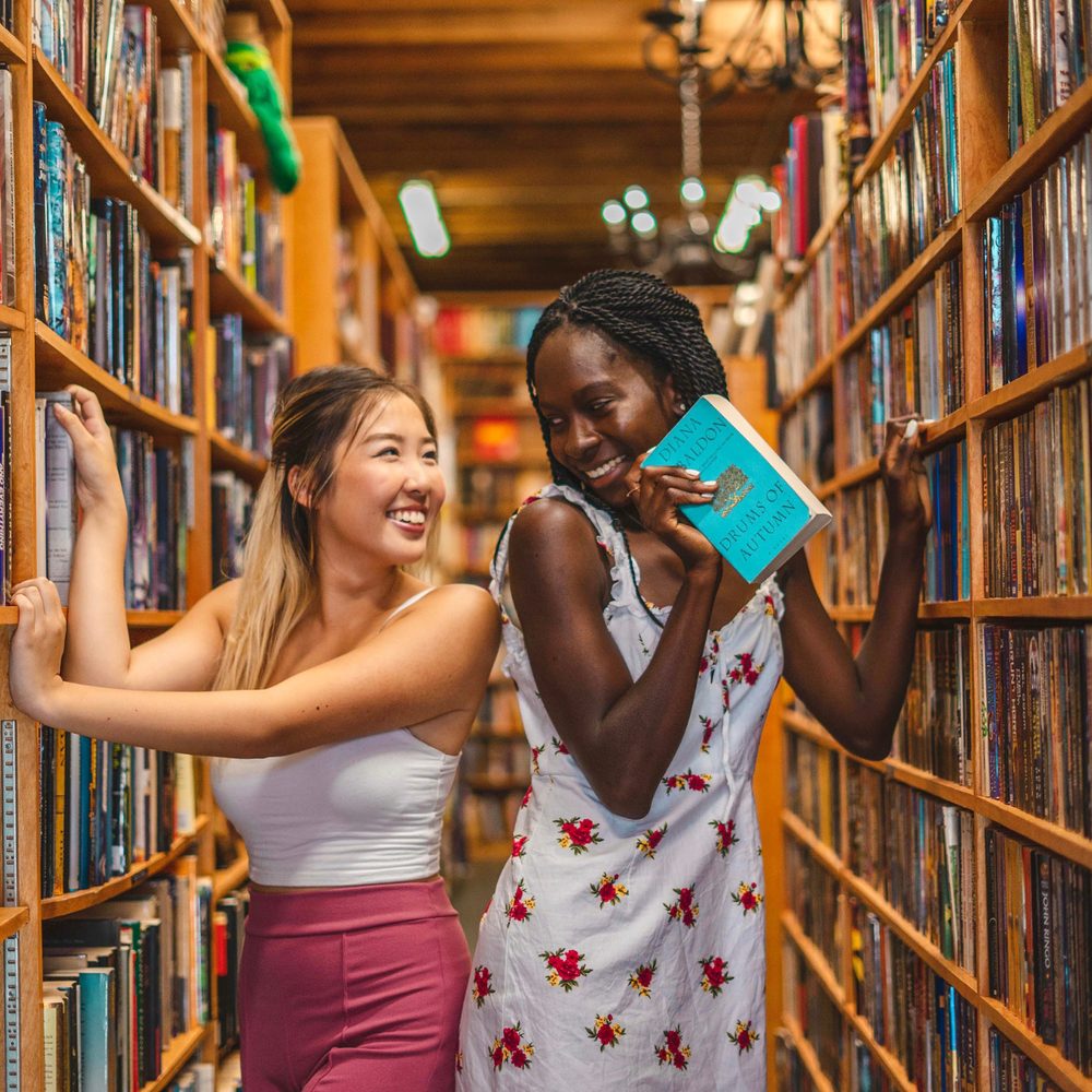 Two smiling students between library shelves, holding a book